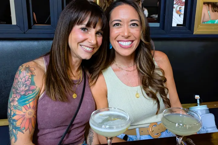 Two women sitting at a table with cocktails, smiling at the camera.