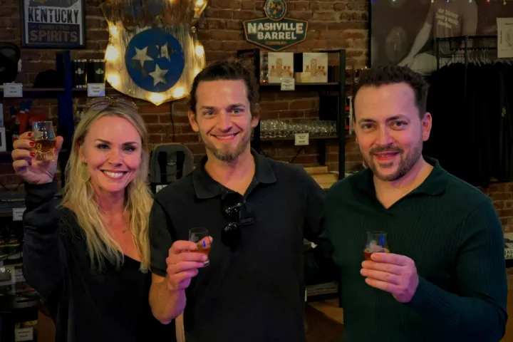 Three people holding drinks, smiling in a bar with Nashville-themed decor.