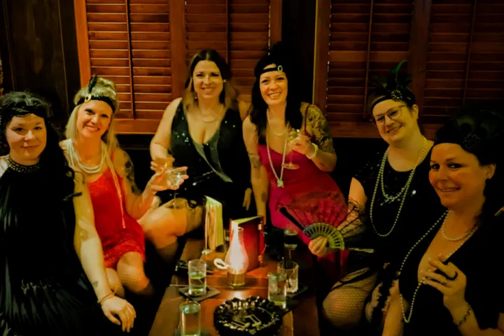 Six women in 1920s flapper attire sit together in a dimly lit room.