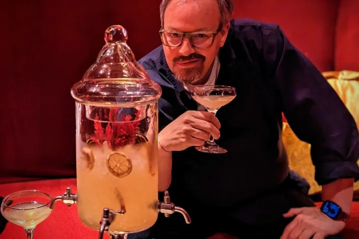 Man holding cocktail beside a drink dispenser with sliced limes, seated on a red couch.