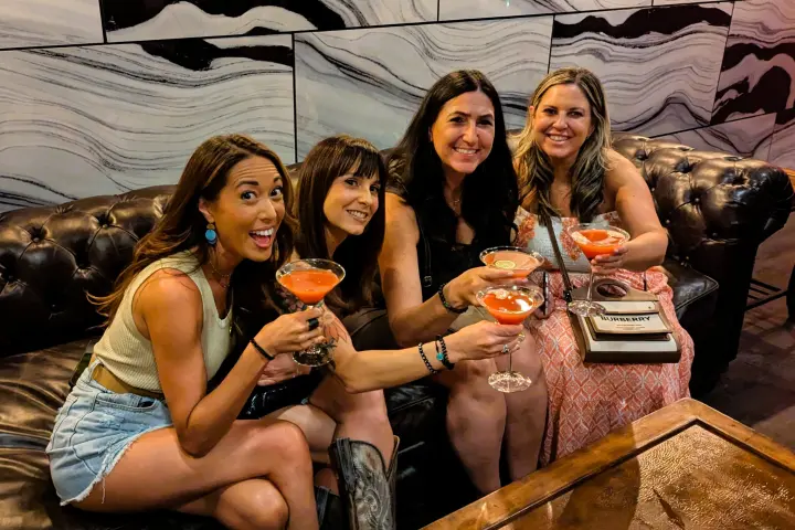 Four women sitting on a couch holding cocktails, smiling at the camera.