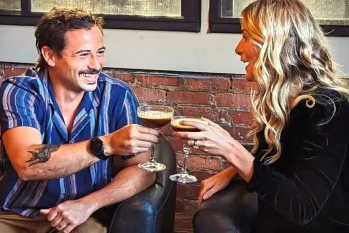 A man and woman smiling while toasting with cocktails in a cozy setting.