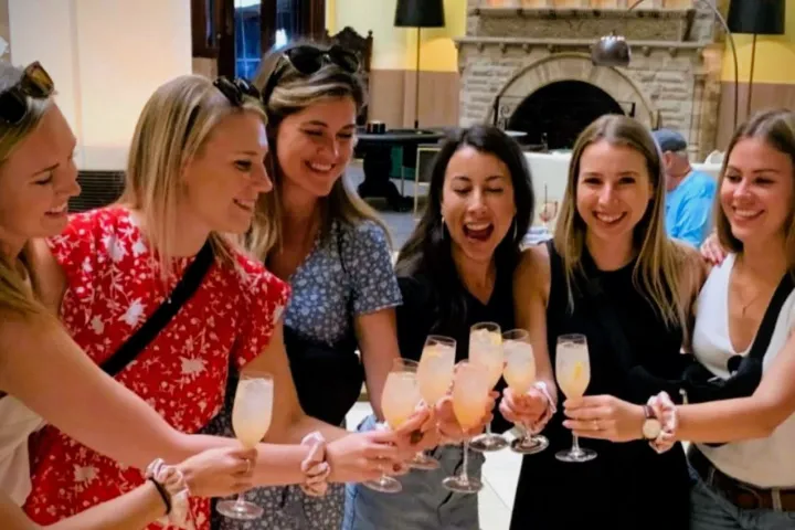 Six women smiling and toasting with drinks indoors.