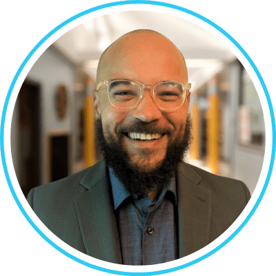 Untitled design (7) Smiling person with glasses and beard, wearing a suit, in a bright indoor setting.